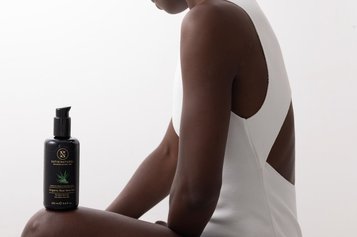 Dunkle Aloe-Vera-Gel-Flasche von Satin Naturel auf dem Knie einer sitzenden Frau in weissem Body — Das Studiozwei, Werbefotograf München