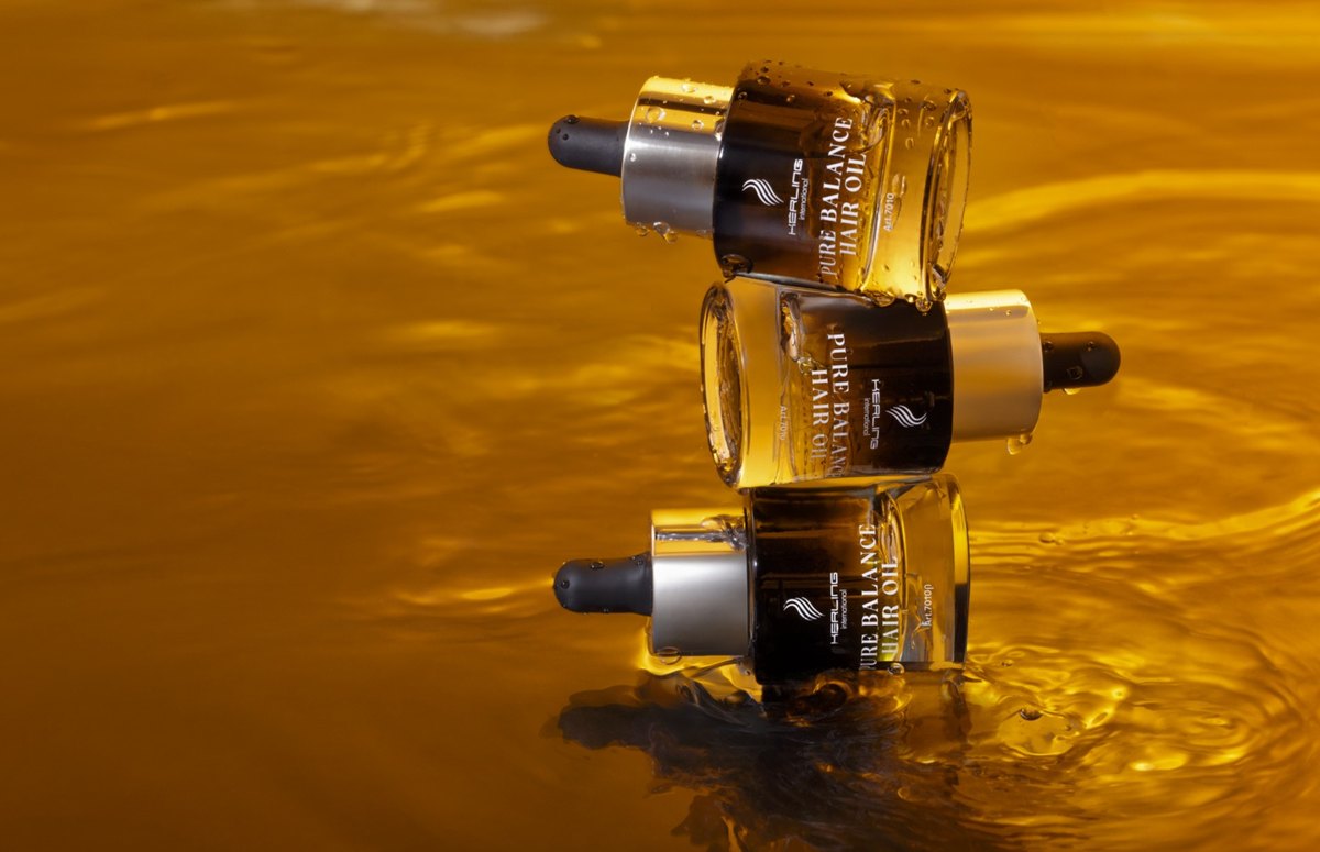 Drei Pure Balance Hair Oil Flaschen liegen gestapelt auf einer goldgelben, bewegten Oelflaeche — Das Studiozwei, Werbefotograf München