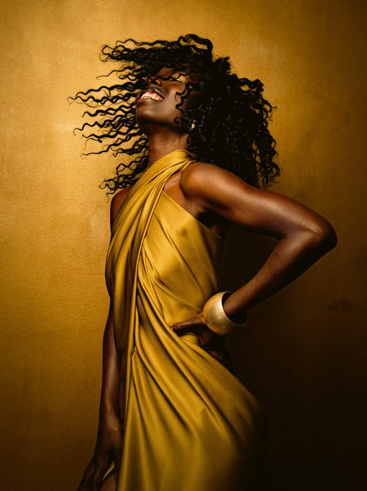 Model in goldgelbem drapiertem Kleid mit fliegendem Haar vor goldbraunem Hintergrund — Das Studiozwei, Werbefotograf München
