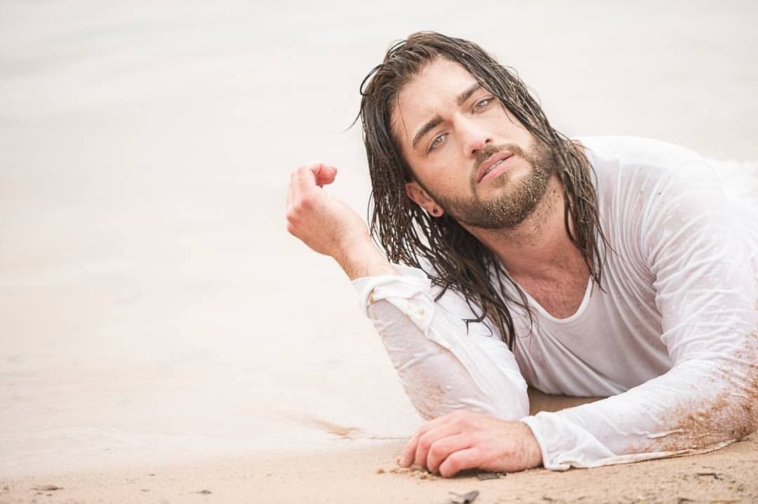 Mann mit nassen langen Haaren und weissem Shirt liegt im hellen Sand am Wasser — Das Studiozwei, Werbefotograf München