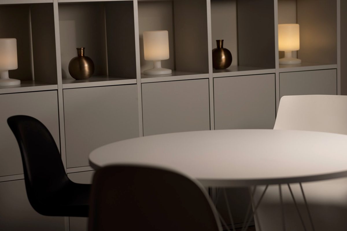 Runder weisser Tisch mit Schalenstuehlen vor einem Regal mit warm leuchtenden Tischlampen — Das Studiozwei, Werbefotograf München