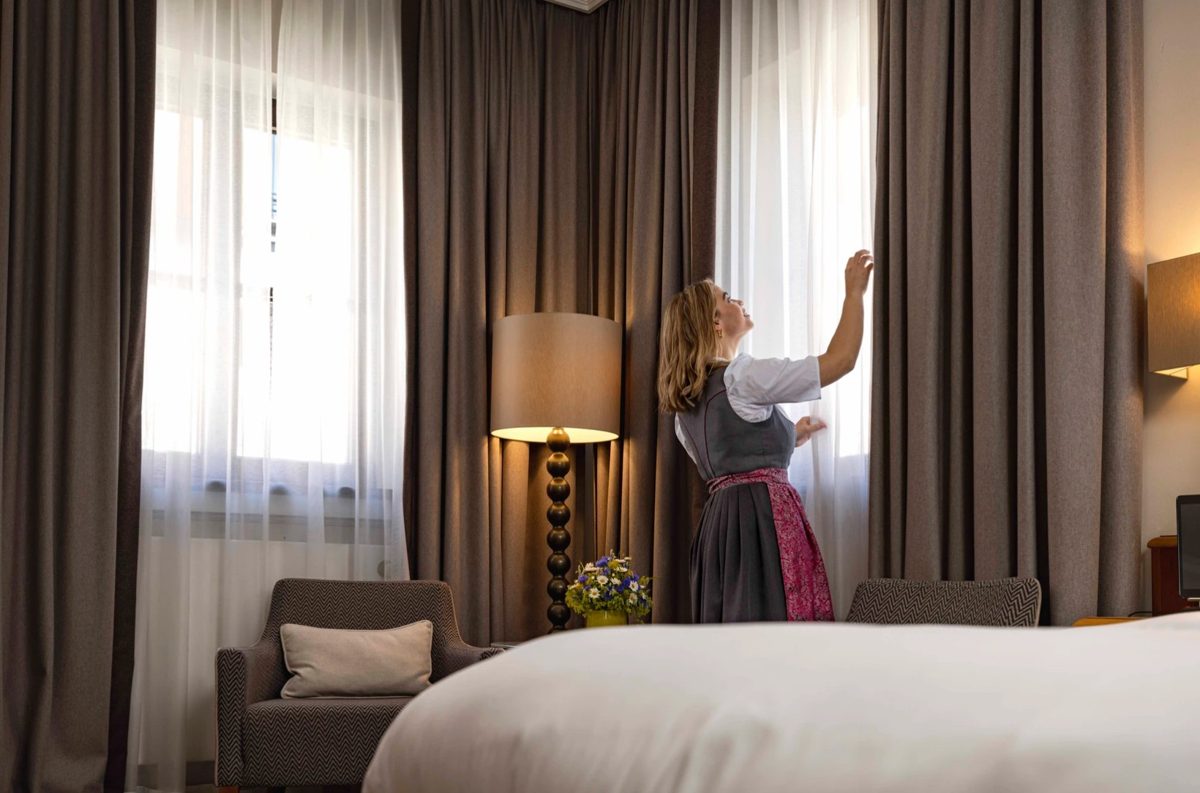 Frau in Dirndl oeffnet den Vorhang in einem Hotelzimmer mit Bett und Sessel — Das Studiozwei, Werbefotograf München