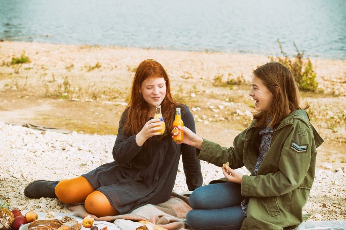 Zwei Freundinnen stossen beim Picknick am See mit Limonadenflaschen an — Das Studiozwei, Werbefotograf München