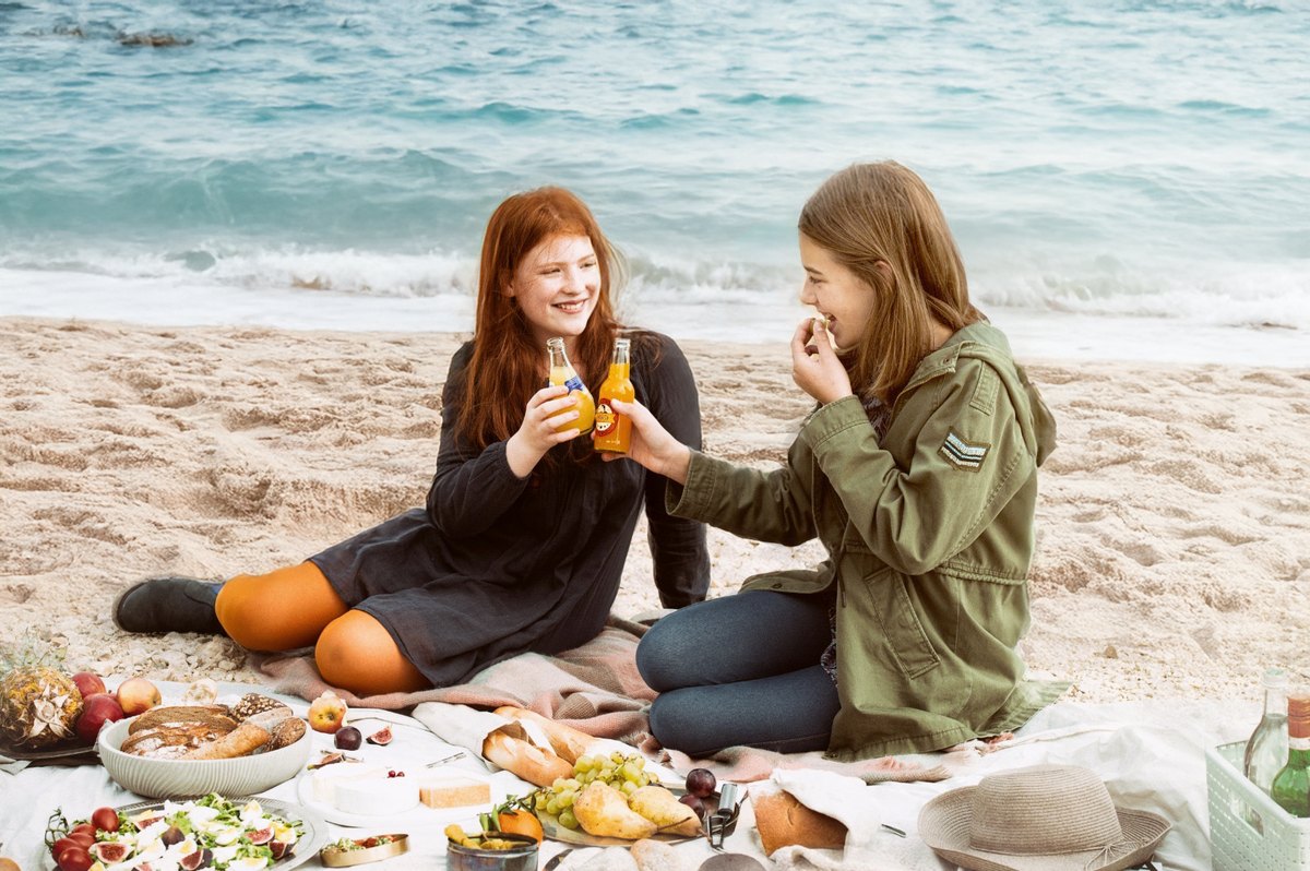 Zwei Frauen beim Picknick am Strand stossen mit Flaschen an — Das Studiozwei, Werbefotograf München