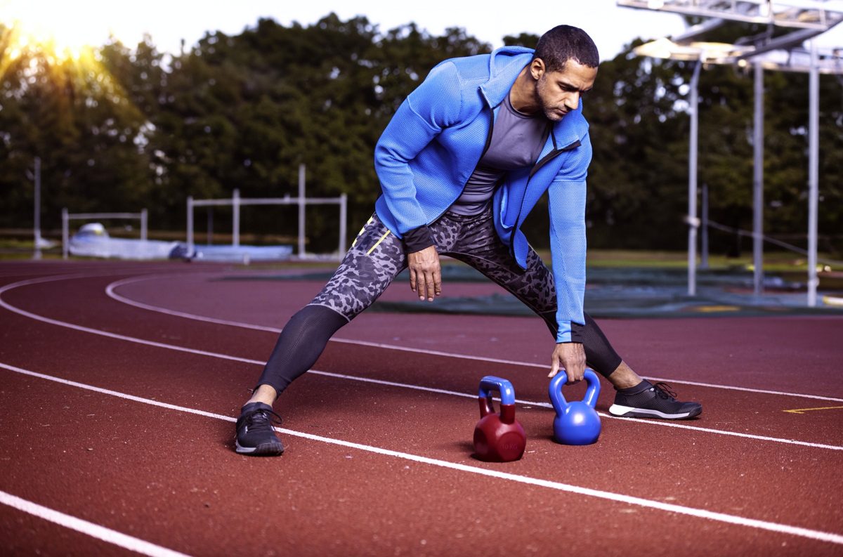 Sportler dehnt sich auf einer roten Laufbahn neben zwei Kettlebells im Gegenlicht — Das Studiozwei, Werbefotograf München