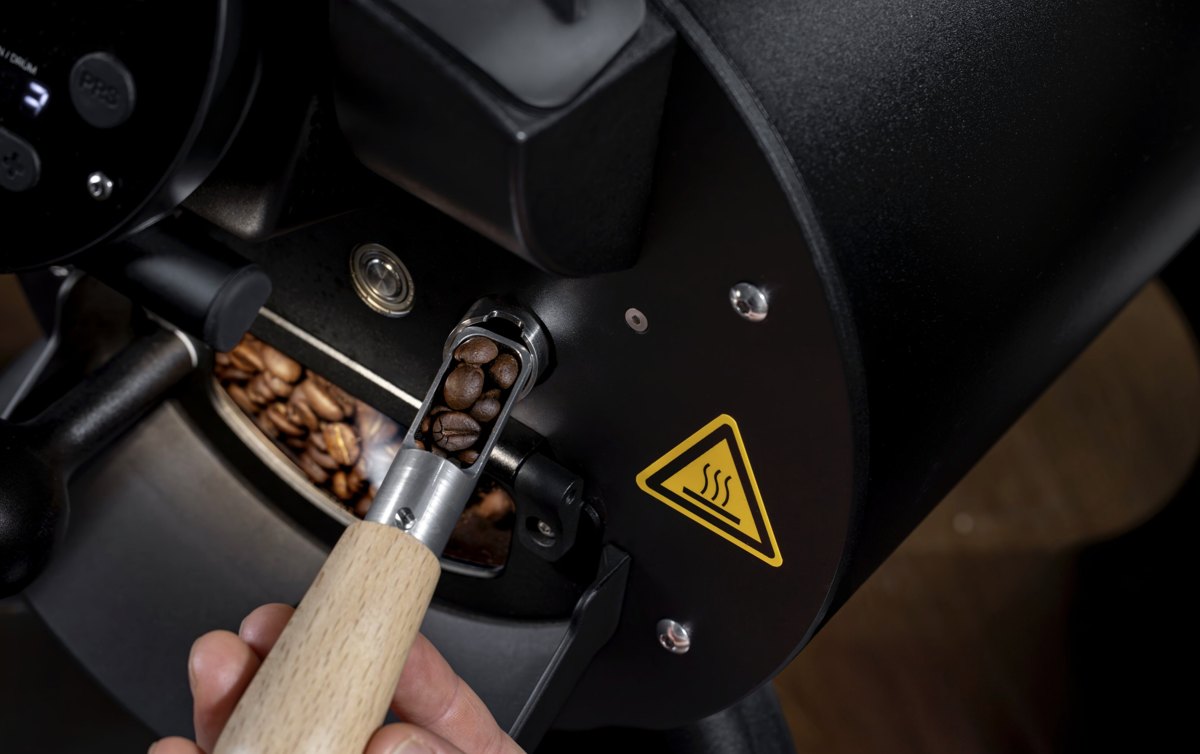 Hand entnimmt mit einem Holzloeffel geroestete Kaffeebohnen aus einem schwarzen Roester — Das Studiozwei, Werbefotograf München