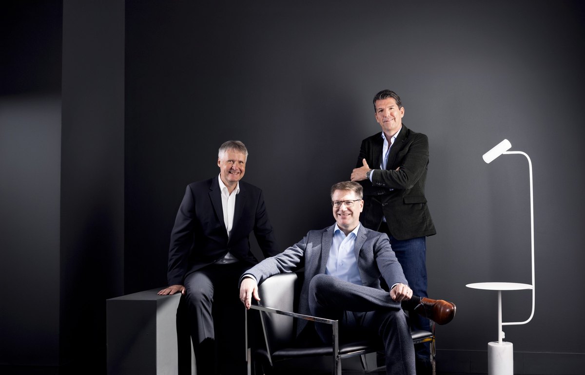 Drei Maenner in Business-Kleidung gruppiert um schwarzen Lederstuhl vor dunklem Hintergrund — Das Studiozwei, Werbefotograf München