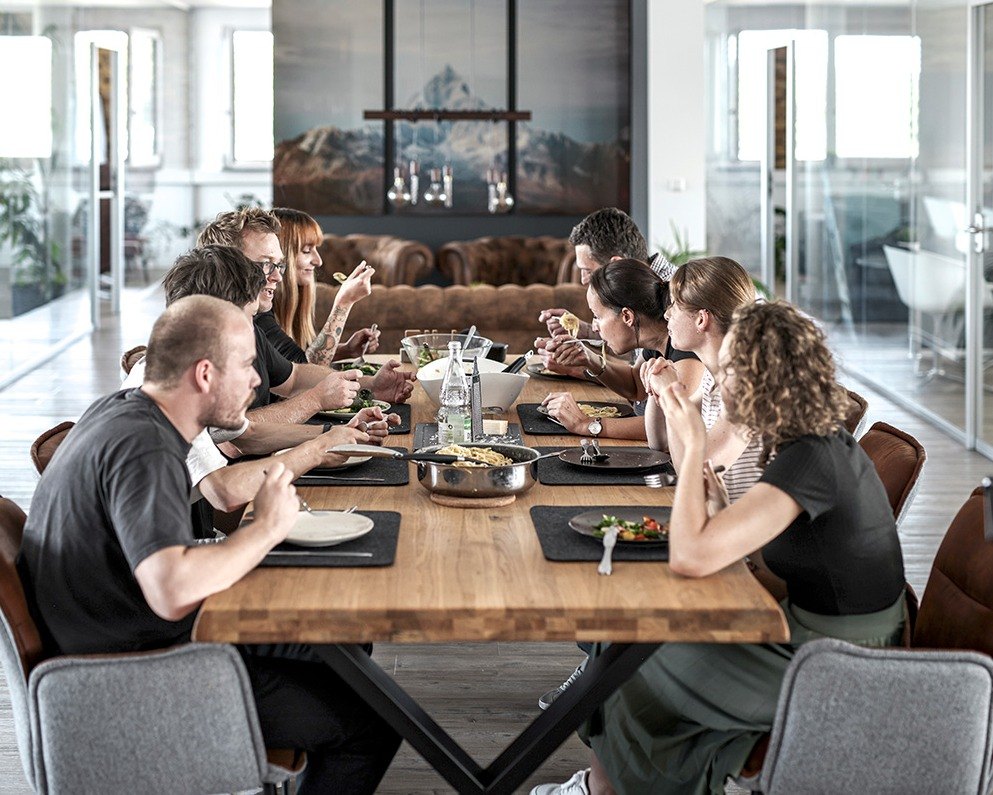 Grosse Gruppe beim gemeinsamen Mittagessen an langer Holztafel im offenen Buero — Das Studiozwei, Werbefotograf München
