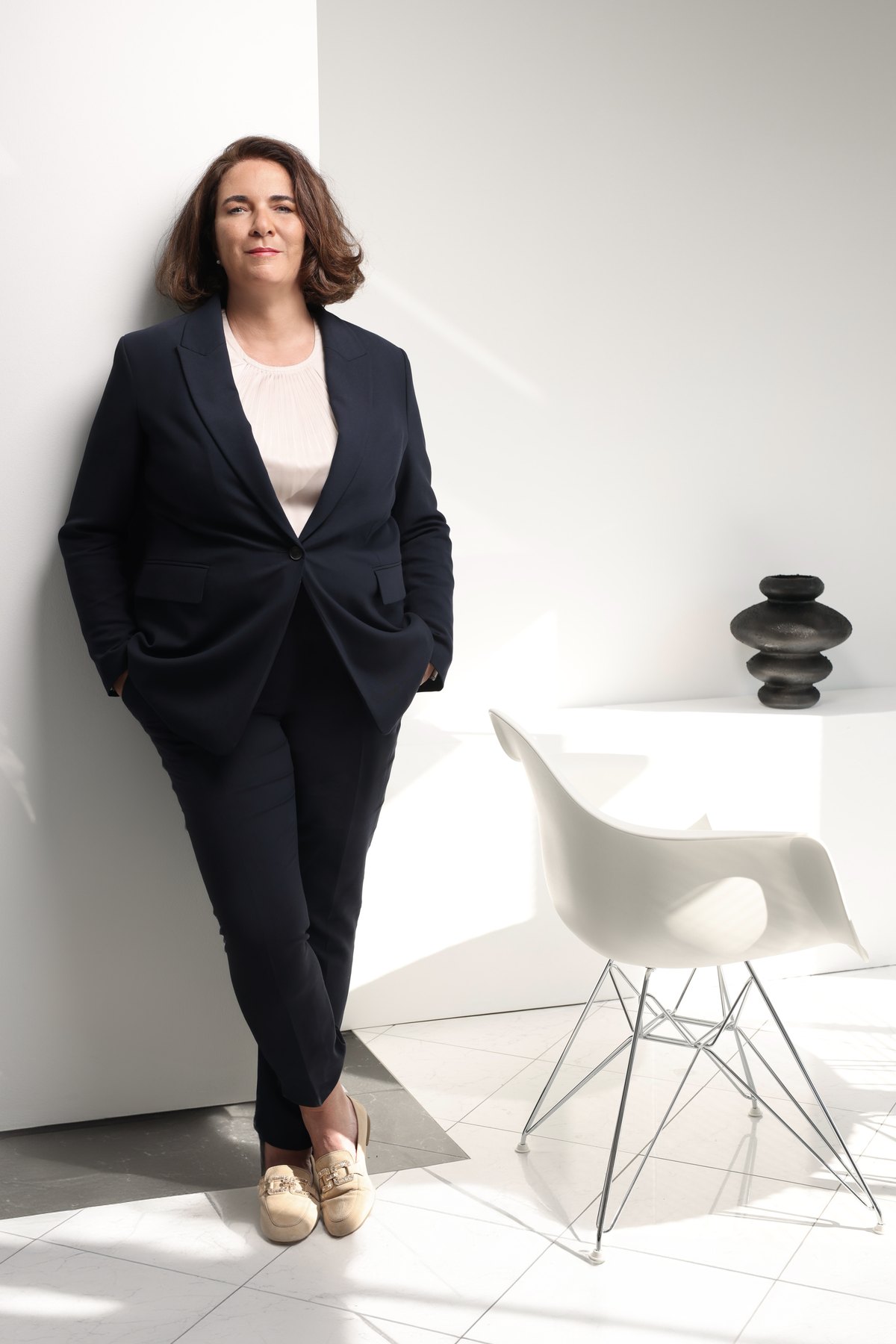 Frau im dunklen Anzug steht in hellem Raum neben Designstuhl — Das Studiozwei, Werbefotograf München