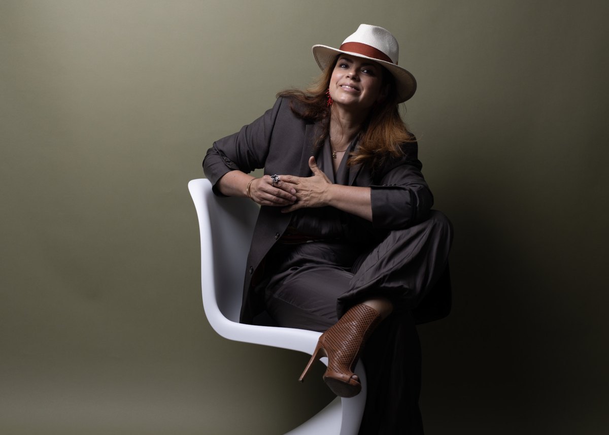 Cristina Galler mit hellem Hut sitzt seitlich auf einem weissen Schalenstuhl vor olivgruener Wand — Das Studiozwei, Werbefotograf München