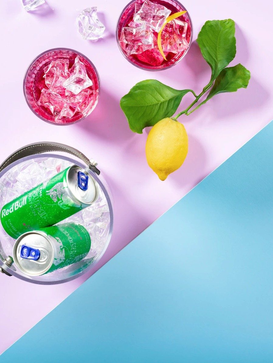 Eiskuebel mit gruenen Dosen, pinke Drinks und Zitrone von oben auf flieder-blauem Hintergrund — Das Studiozwei, Werbefotograf München