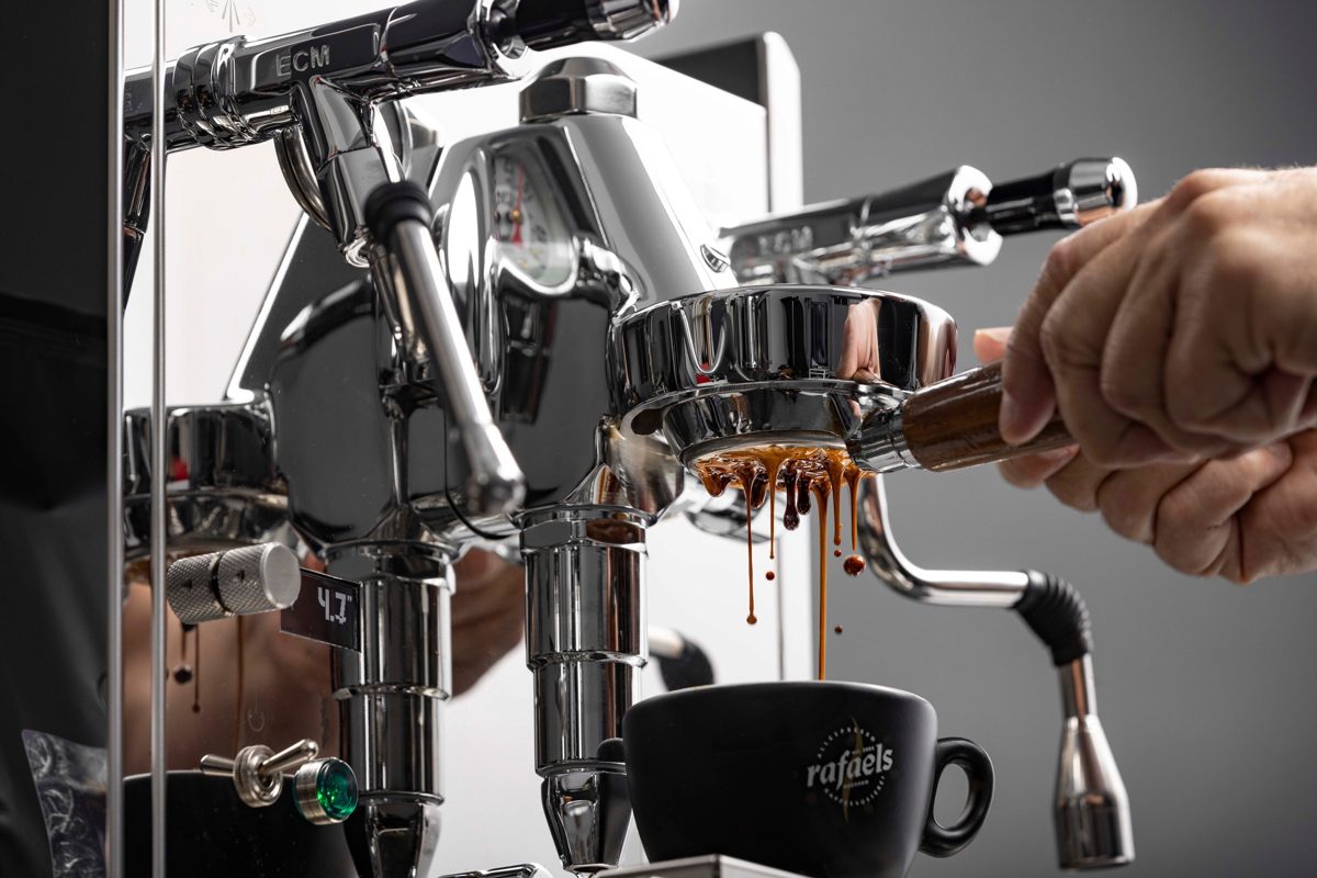 Nahaufnahme einer verchromten Espressomaschine, aus der Kaffee in eine schwarze Tasse laeuft — Das Studiozwei, Werbefotograf München