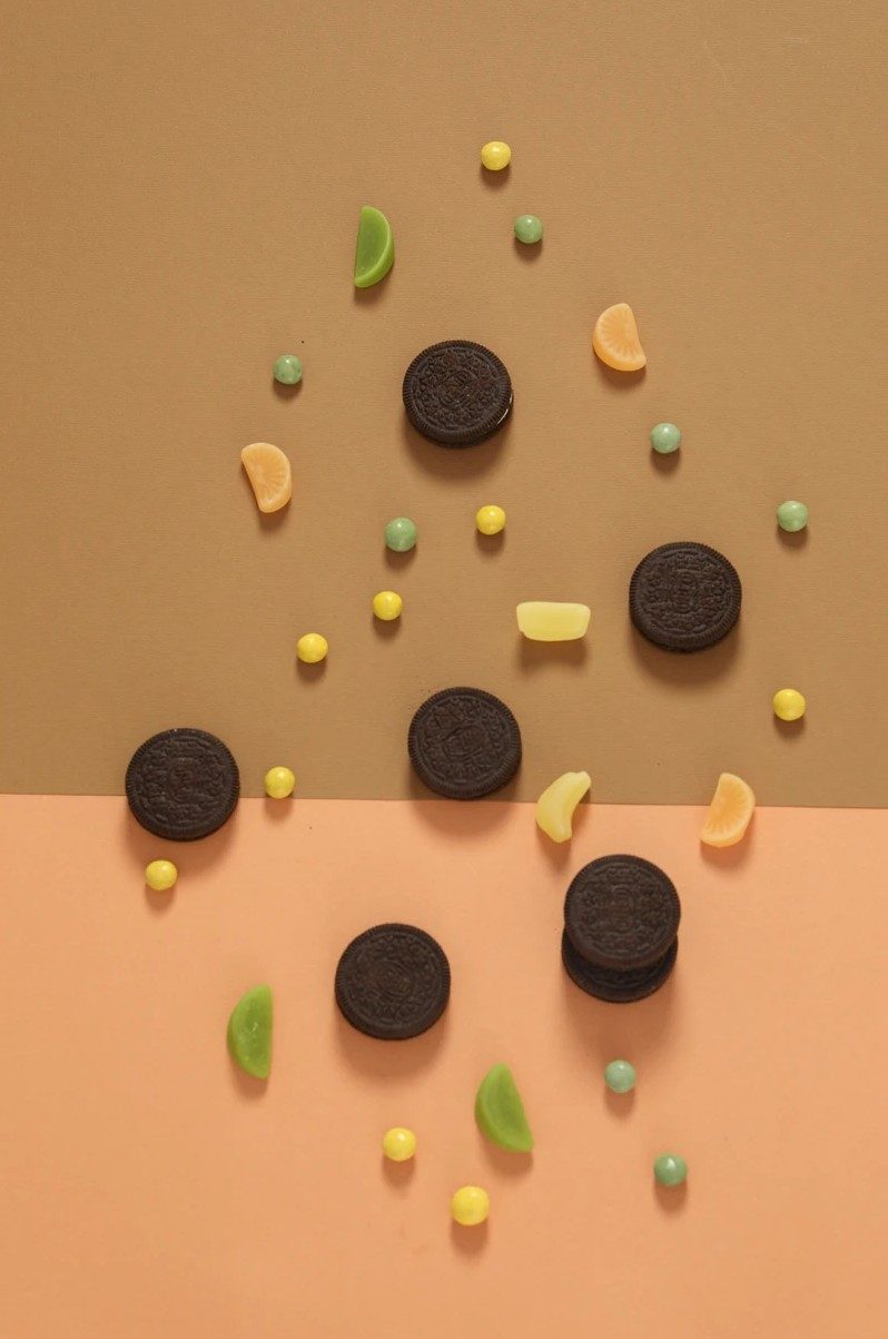 Dunkle runde Kekse und bunte Bonbons verteilt auf oliv-apricotfarbenem Hintergrund — Das Studiozwei, Werbefotograf München