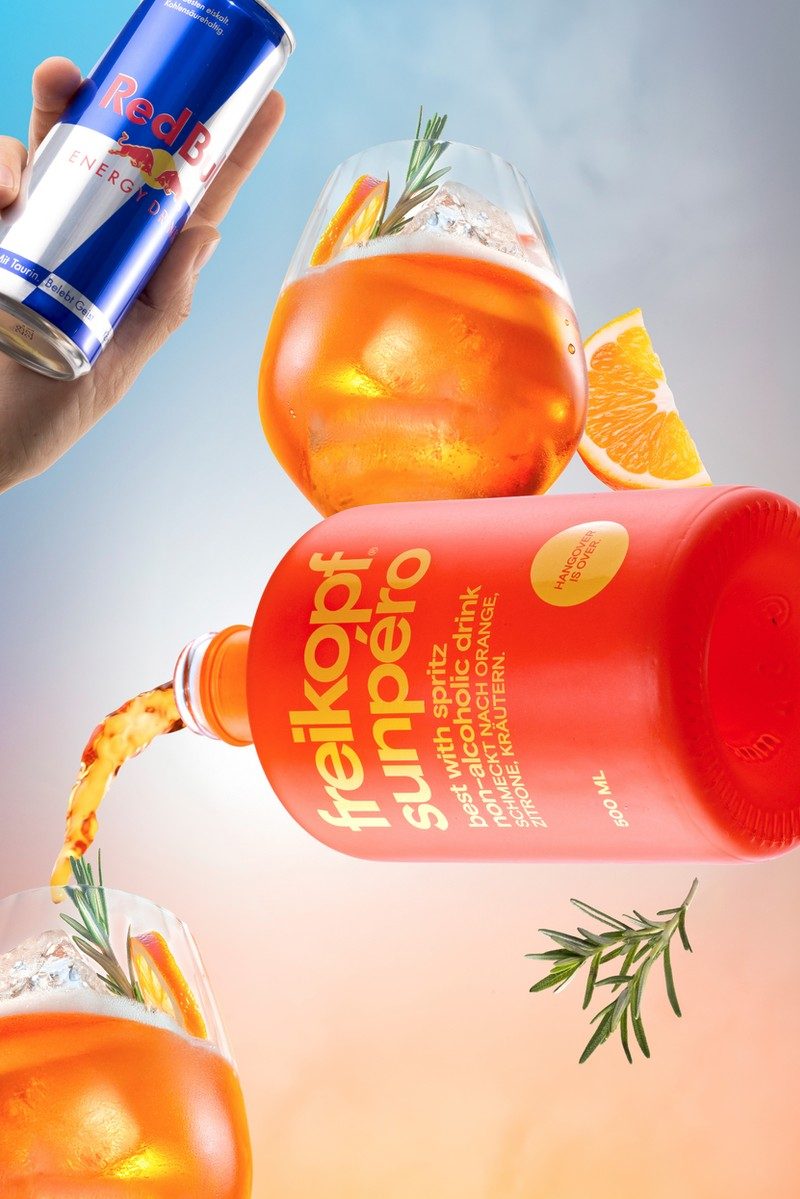 Schwebende orangefarbene Flasche mit Spritz-Glas, Orangenscheiben und Rosmarin vor blau-pfirsichfarbenem Verlauf — Das Studiozwei, Werbefotograf München