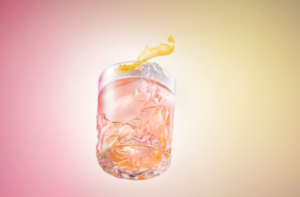 Schwebendes Kristallglas mit rosa Drink, Eis und Zitronenzeste vor pink-gelbem Verlauf — Das Studiozwei, Werbefotograf München