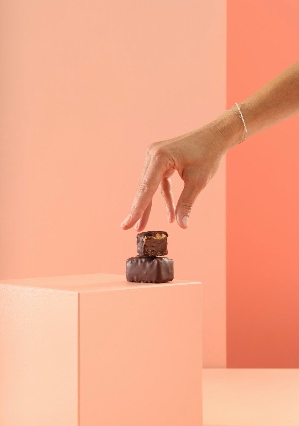 Hand greift nach gestapelten Schokoladenstuecken auf apricotfarbenem Podest im Studio — Das Studiozwei, Werbefotograf München