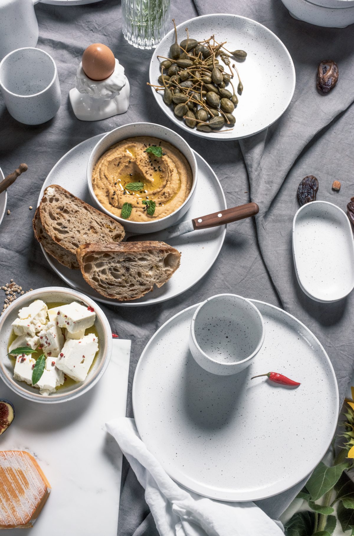 Aufsicht auf gesprenkeltes weisses Geschirr mit Hummus, Brot, Feta und Kapern auf grauem Leinen — Das Studiozwei, Werbefotograf München